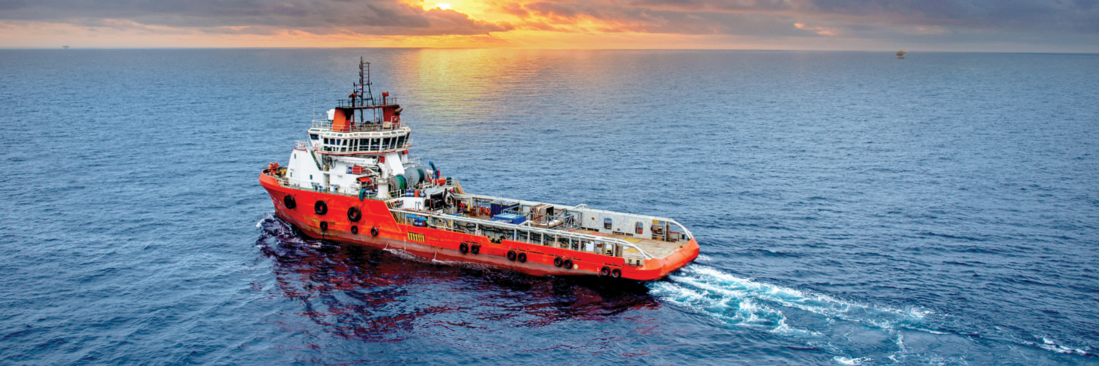 Forsyningsskip i rød farge på åpent hav under solnedgang, brukt til offshore olje- og gassoperasjoner. Skipet lager kjølvann mens det beveger seg mot horisonten.