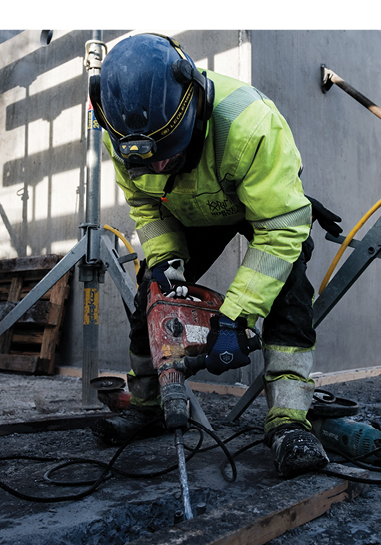 Bygningsarbeider i verneklær og hjelm bruker kraftig boremaskin på betong, med slitesterke arbeidshansker for beskyttelse.