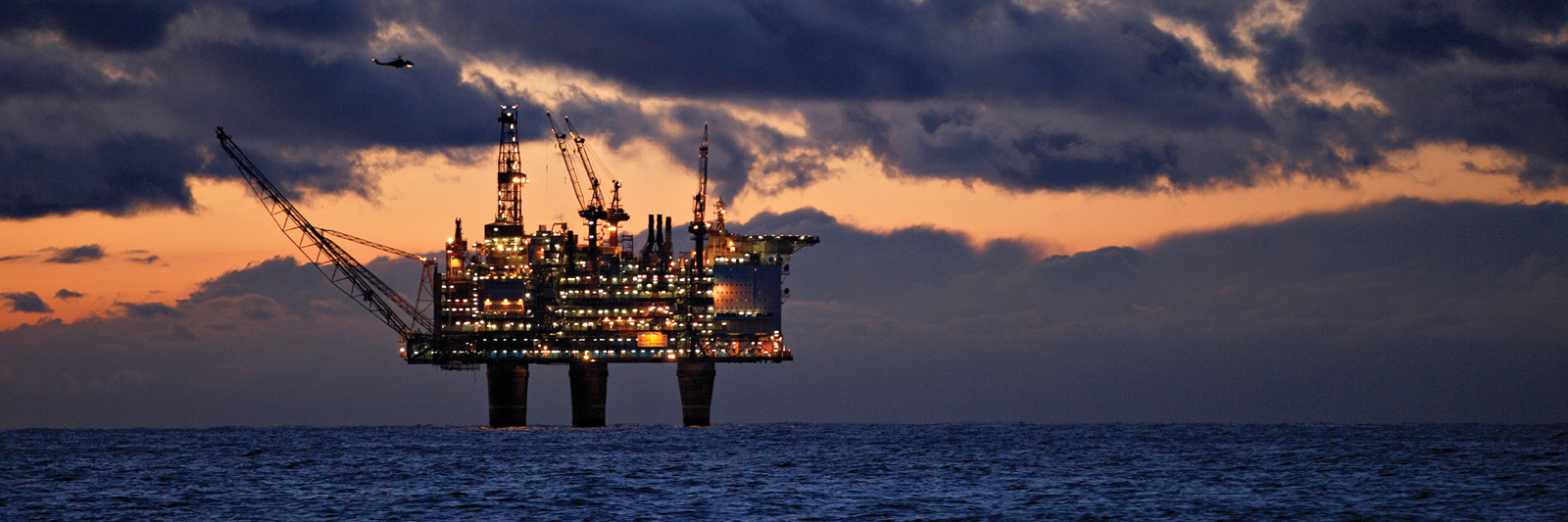 Opplyst offshore oljeplattform med kraner og strukturer i Nordsjøen ved solnedgang, med helikopter i luften og mørke skyer i bakgrunnen.